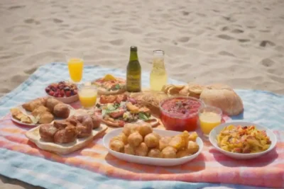 Una amplia variedad de platos gourmet y bebidas en una manta de picnic de playa bajo el sol.